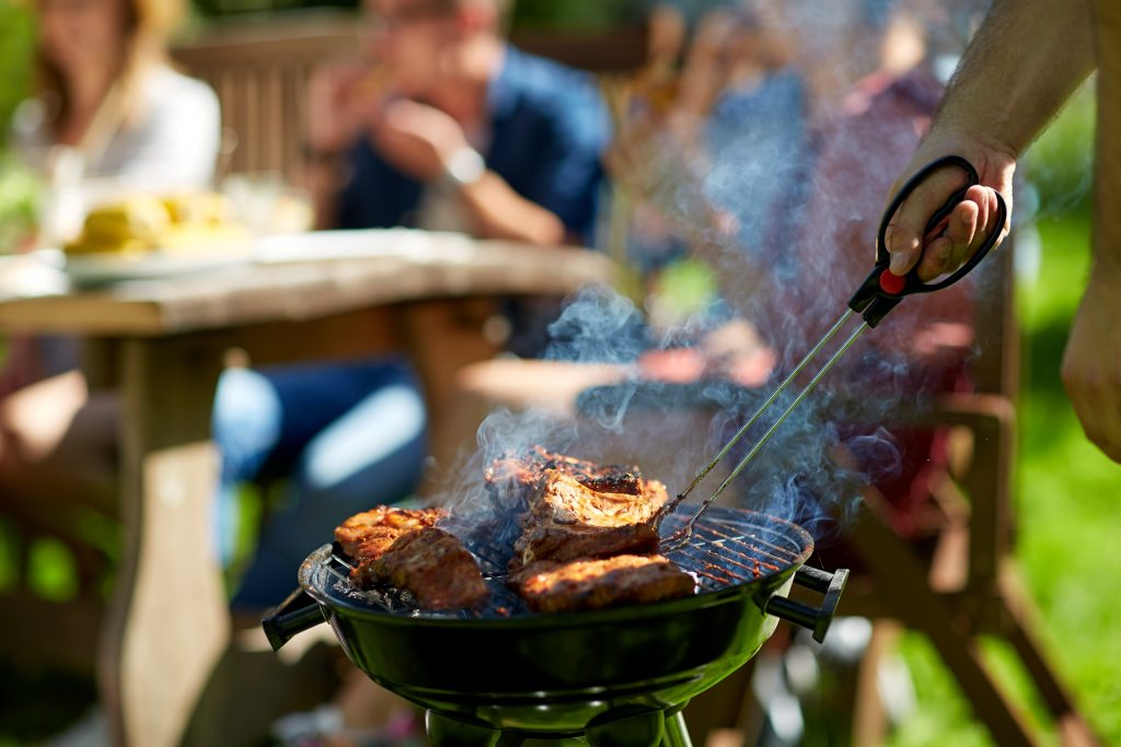 leisure, food, people and holidays concept - man cooking meat on barbecue grill for his friends at summer outdoor party