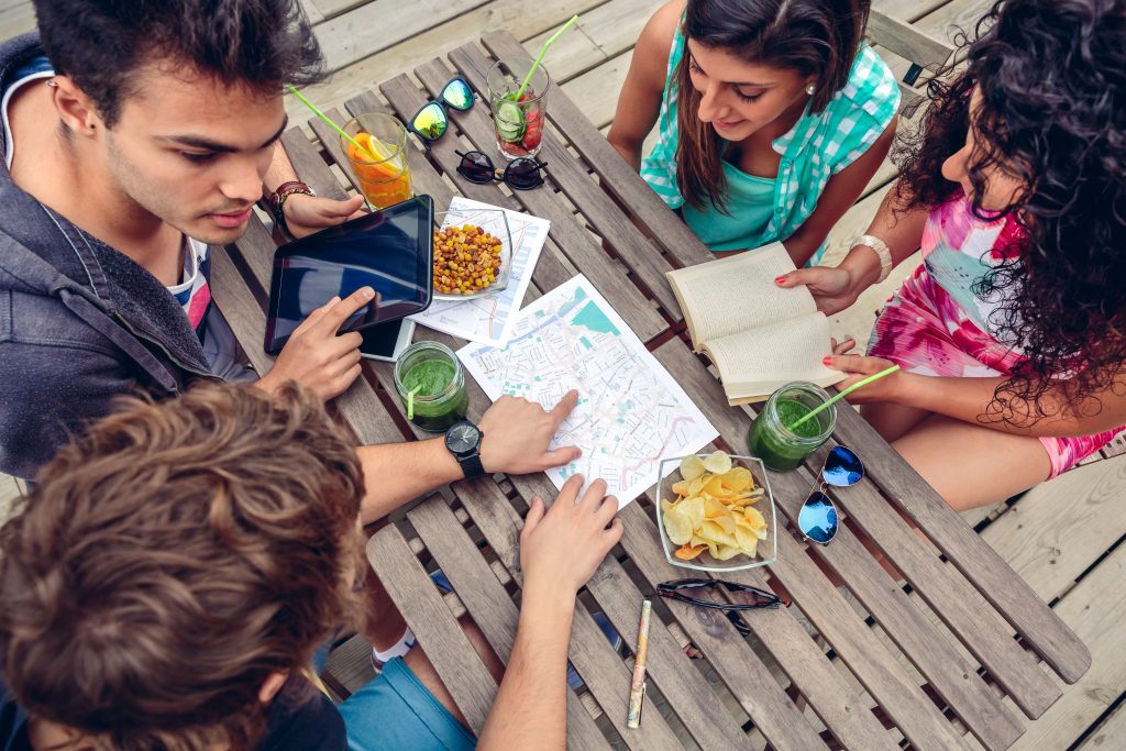 Young friends looking map over a wooden table with healthy drinks and snacks. Holidays and tourism concept.