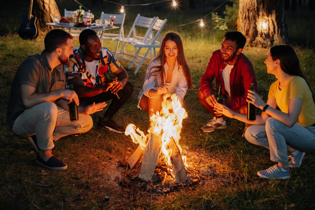 Group of friends sitting around bonfire, talking, drinking beer and enjoying summer night outdoors