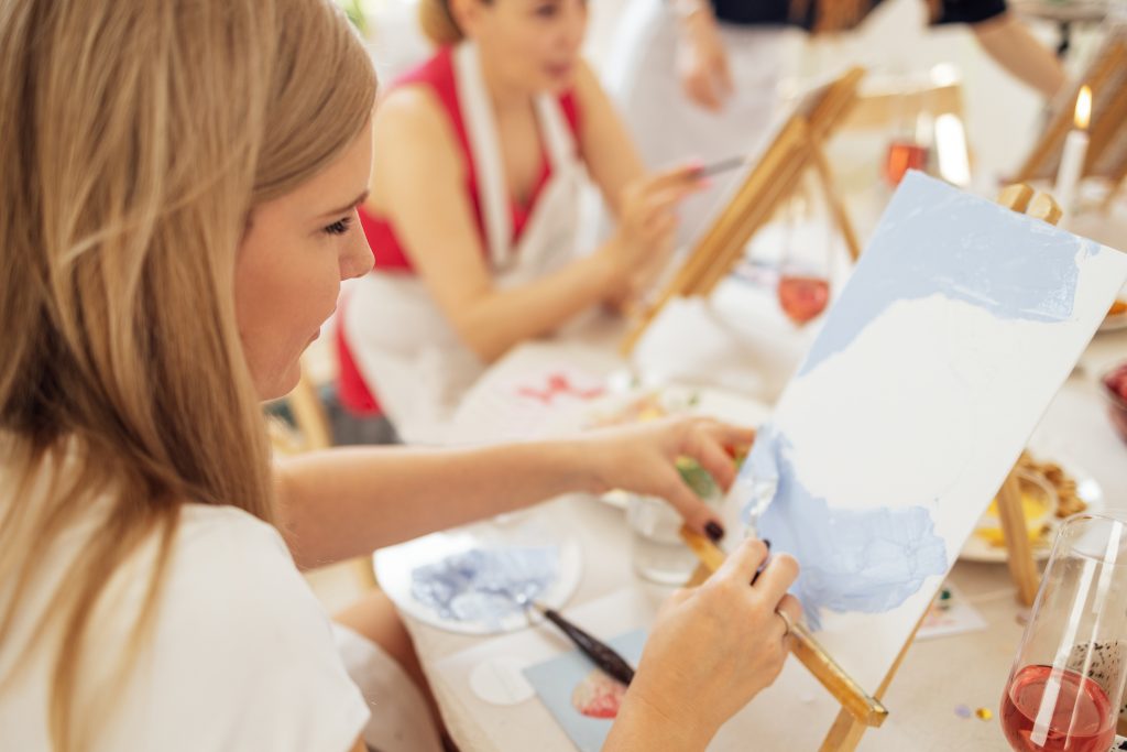 Close-up of canvas on easel and hand with spatula. Young artist paints picture at table. Blonde in painting workshop. Girl is learning to draw in art studio. Creative activity and hobby