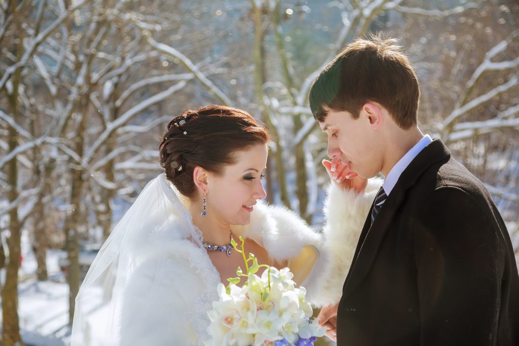 Bride groom stand together in winter wonderland, surrounded by snow covered trees bride holds bouquet of white flowers, they share tender moment.