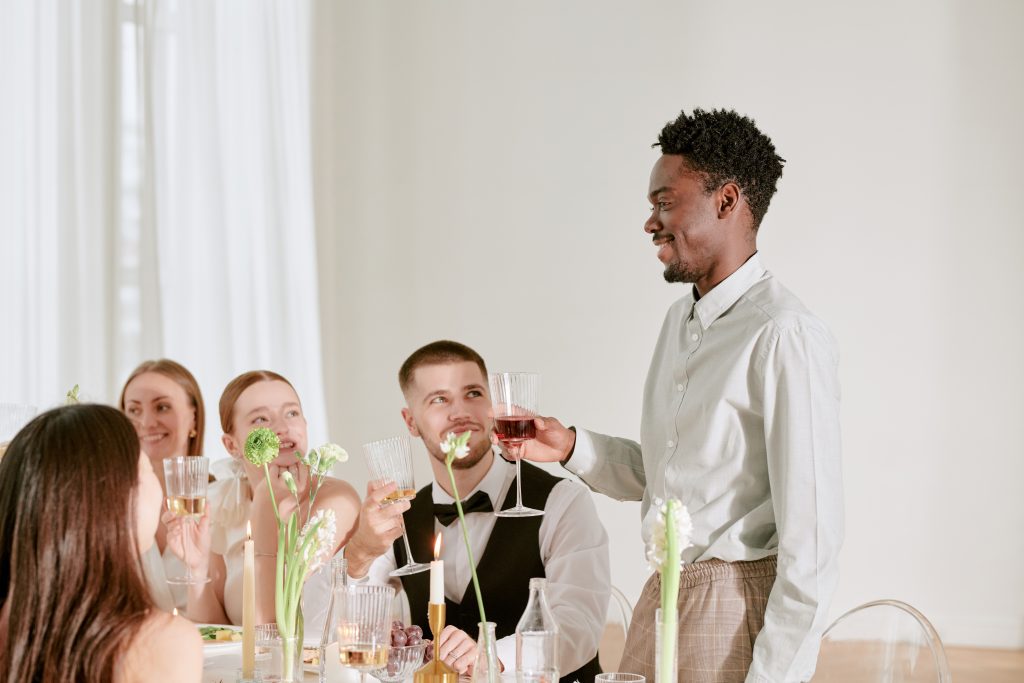 Group of young adult multiethnic men and women sitting at wedding celebration table smiling and raising glasses while man standing and giving toast